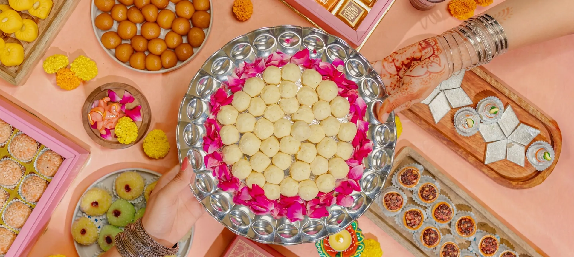 Hands serving a silver thali of laddoos surrounded by festive sweets
