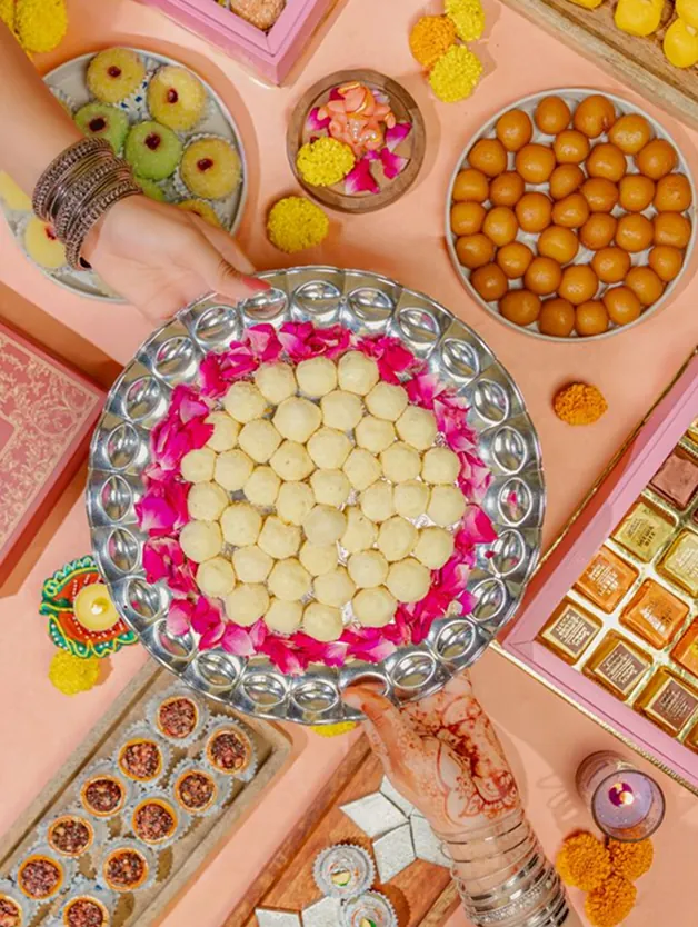 Hands serving a silver thali of laddoos surrounded by festive sweets
